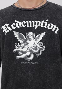 Svart, oversized t-skjorte med en teksturert finish. Har et grafikk av en engel som holder en slange og teksten "Redemption" i fet hvit.