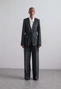 Szürke öltöny, övvel ellátott blézerrel, fehér varrással és széles szárú nadrággal, fehér inggel párosítva. Simára bontott szövettextúra.