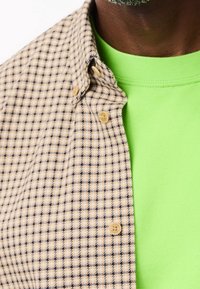 Beżowa koszula w kratę z ciemnoniebieskimi konturami, z okrągłym kołnierzykiem i jasnymi guzikami, zestawiona z jaskrawą zieloną bluzą z okrągłym dekoltem.