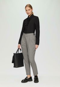 Frau steht und trägt eine schwarze Bluse mit langen Ärmeln, graue High-Waist-Hosen, schwarze Loafers und hält eine schwarze Tragetasche.