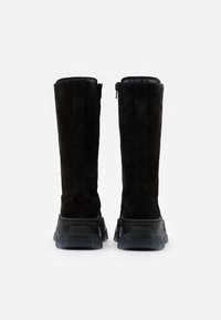 Para wysokich czarnych zamszowych botków z grubymi, teksturowanymi podeszwami, widocznych od tyłu na białym tle.