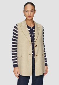 Beige Wollweste mit maßgeschneidertem Design, einreihigem Stil, zwei vorderen Taschen und braunen Knöpfen, getragen über einem gestreiften Pullover.
