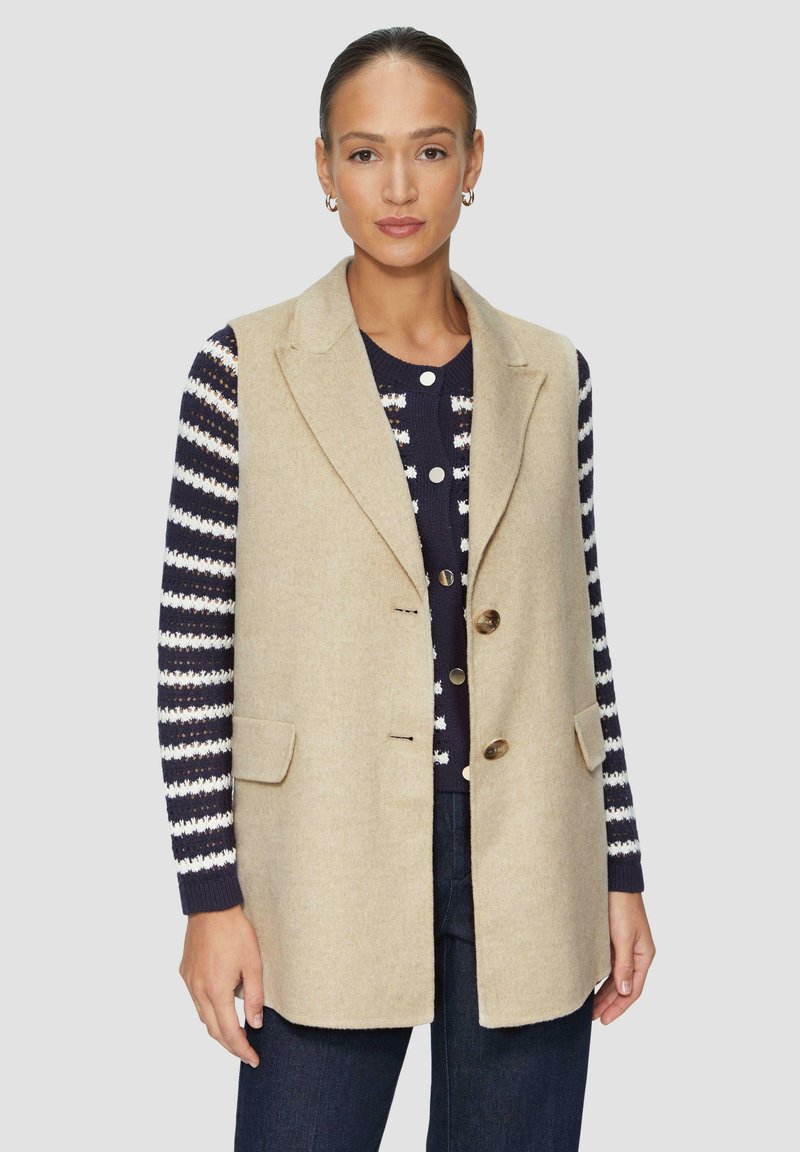 Beige Wollweste mit maßgeschneidertem Design, einreihigem Stil, zwei vorderen Taschen und braunen Knöpfen, getragen über einem gestreiften Pullover.