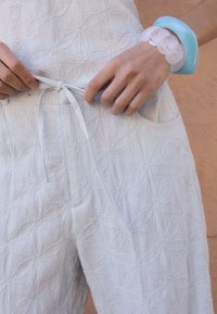 Světle modrá textilní látka s texturou a vzorem listů. Má stahovací pás a viditelné boční kapsy. Náramek vyrobený z průhledného a světle modrého plastu.