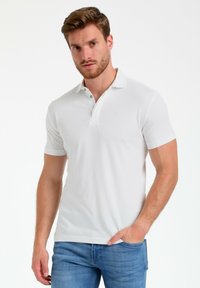 Camisa polo blanca de manga corta con una tapeta de dos botones, cuello plano y un sutil detalle de logo. Combinada con jeans de color azul claro.