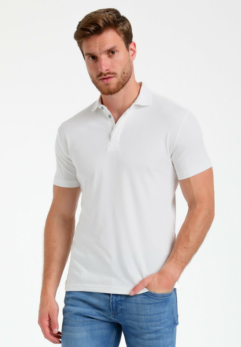 Camisa polo blanca de manga corta con una tapeta de dos botones, cuello plano y un sutil detalle de logo. Combinada con jeans de color azul claro.