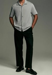 Camicetta grigia a maniche corte con collo e bottoni, abbinata a pantaloni neri larghi e scarpe nere. Design semplice con texture liscia.