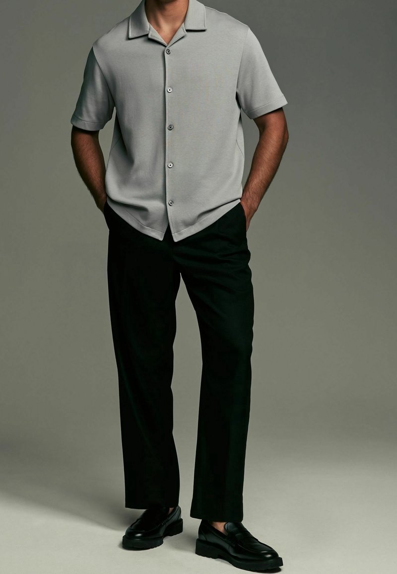 Camicetta grigia a maniche corte con collo e bottoni, abbinata a pantaloni neri larghi e scarpe nere. Design semplice con texture liscia.