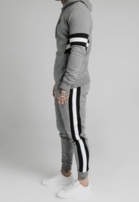 Mężczyzna w szarej bluzie z kapturem i dresowych spodniach z czarno-białymi paskami na rękawach i nogawkach, w białych trampkach, stojący bokiem na szarym tle.