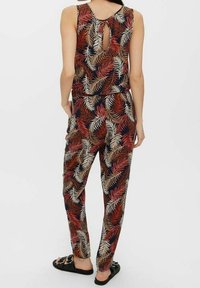Combinaison multicolore avec un motif de feuilles dans des teintes de rouge, beige et marine. Design avec dos en trou de serrure et coupe décontractée. Portée avec des sandales.