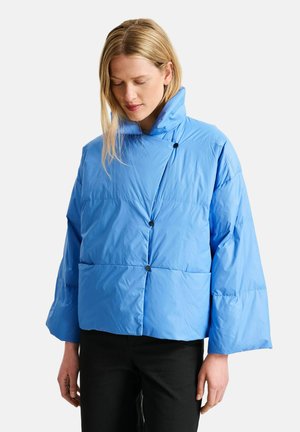 Mujer que lleva una chaqueta acolchada azul claro con cuello alto y cierre asimétrico de botones, combinada con pantalones negros, de pie sobre un fondo blanco.
