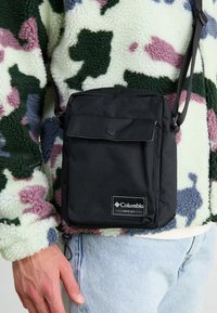 Borsa a tracolla nera in tessuto con chiusura a patta e tasca frontale, caratterizzata da un badge del logo Columbia e una tracolla regolabile.
