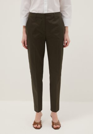 Mujer vistiendo pantalones tobilleros sastre en oliva oscuro, camisa blanca abotonada y sandalias marrones de tacón abiertas, de pie frente a un fondo liso.