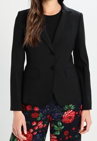 Zwarte getailleerde blazer van een gladde stof, met een reverskraag, twee knopen en voorzakken. Draagt over bloemenpatroon broek.