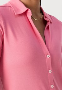 Personne portant une chemise rose vif à boutons avec col et un simple collier chaîne en or sur un fond neutre.