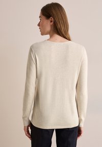 Beige svetr s dlouhými rukávy a měkkou texturou, s výstřihem do lodi a žebrovanými manžetami. Zadní část je mírně delší než přední.