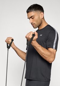 Μαύρο αθλητικό t-shirt με στρογγυλή λαιμόκοψη και κοντά μανίκια, με λευκές ρίγες στα πλάγια. Το πρόσωπο κρατά λαστιχένιες ταινίες άσκησης σε στάση προπόνησης.