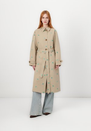 Beige trenchcoat met groene bloemembroideries, knoopsluitingen en een riem. Voorzien van brede mouwen en een split aan de onderkant. Graag gedragen over flared jeans.