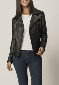 Svart läderjacka med en figursydd design, asymmetrisk dragkedja och breda slag, kombinerad med en vit skjorta och blå jeans.