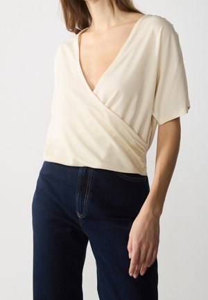 Vrouw draagt een crèmekleurige wikkelblouse met korte mouwen en donkerblauwe, hooggetailleerde jeans tegen een effen witte achtergrond.