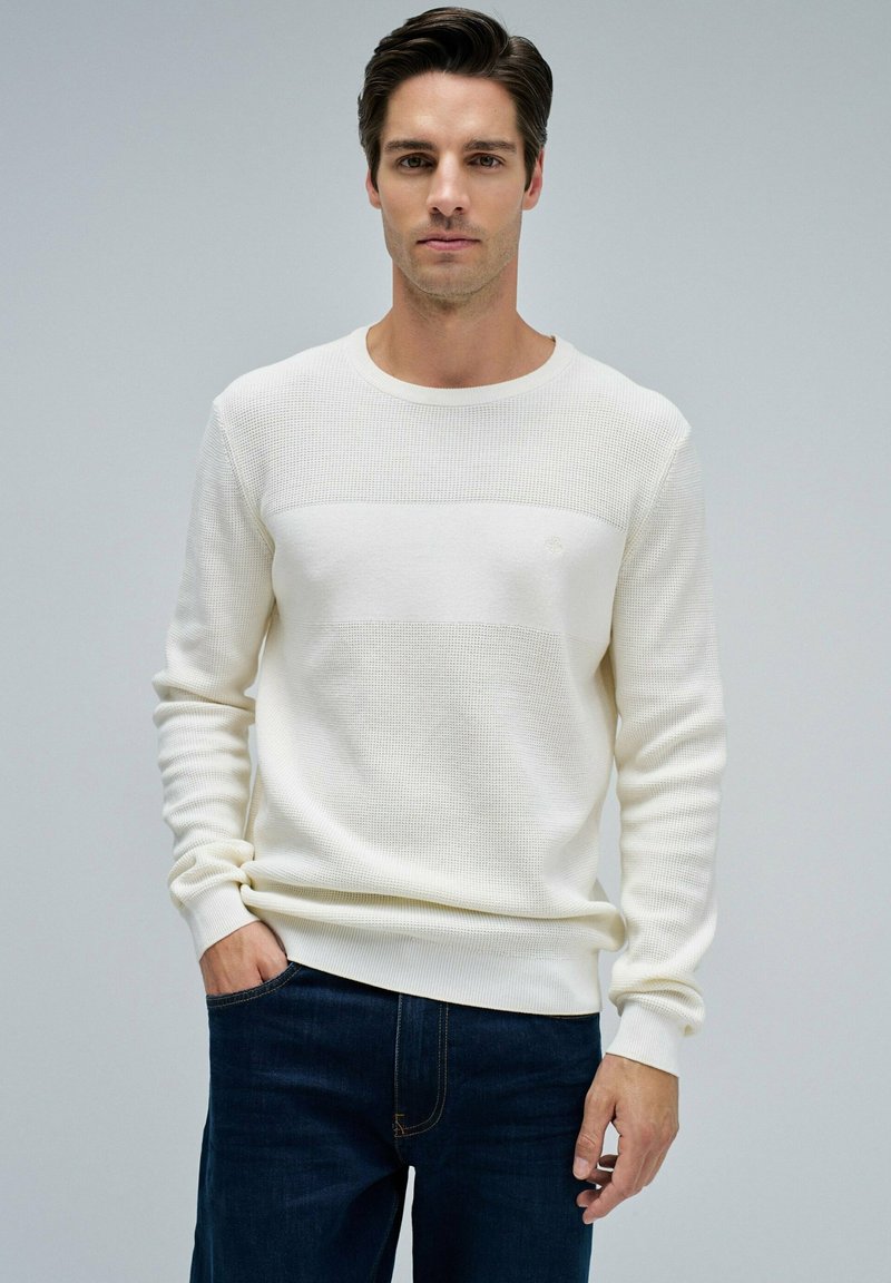 Mann in einem strukturierten cremefarbenen Pullover und dunkelblauen Jeans, der mit einer Hand in der Tasche vor einem schlichten grauen Hintergrund steht.
