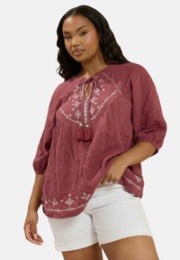 Bordeaux gekleurde blouse met textuur, driekwart mouwen, striknek en witte bloemenborduursels. Gecombineerd met witte shorts.