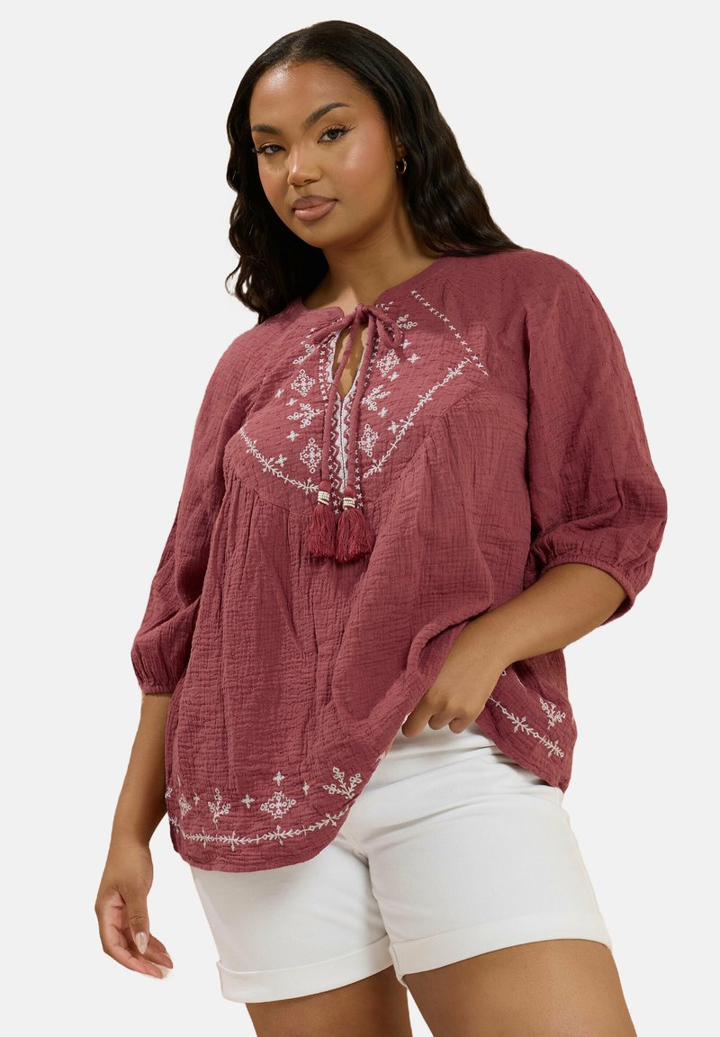 Bordeaux gekleurde blouse met textuur, driekwart mouwen, striknek en witte bloemenborduursels. Gecombineerd met witte shorts.