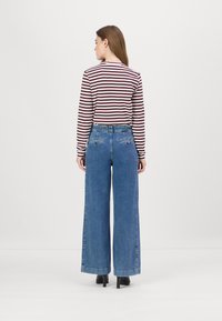 Pantalones vaqueros de pierna ancha en azul medio, con una cintura alta, bolsillos traseros y un sutil logotipo bordado. Combinados con una camiseta de manga larga a rayas.