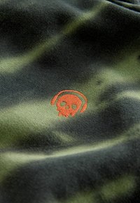 Grüner Camouflage-Stoff mit strukturierter Oberfläche, versehen mit einem orangefarbenen gestickten Totenkopf-Logo mit Kopfhörern, platziert auf der linken Seite.