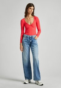 Maglietta rossa a maniche lunghe con scollatura profonda e dettaglio arricciato, abbinata a jeans a gamba larga di colore blu chiaro. Sneakers bianche completano il look.