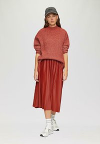 Rode geribbelde trui met hoge hals, gecombineerd met een geplooide rode midi rok. Model draagt een grijze pet en witte sneakers met gestreepte sokken.