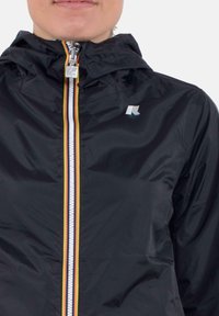 Giacca leggera nera con cappuccio, dotata di una cerniera frontale con strisce colorate e un logo sul petto. Tessuto liscio e lucido.