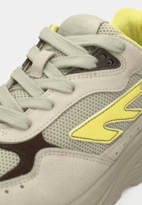 Svetlo siva tenisica s mrežastim panelima, žutim naglascima, smeđim detaljima i teksturiranim potplatom. Ima ravne vezice i podstavljeni ovratnik.