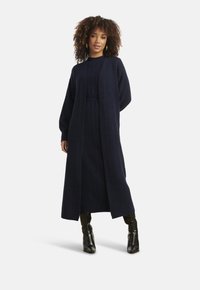 Donna con capelli ricci che indossa un lungo cardigan blu navy e un vestito abbinato a stivali neri al ginocchio, in piedi con una mano dietro la schiena.