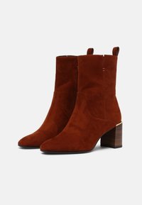 Bottes cheville en daim d'un riche brun rougeâtre avec un bout pointu et un talon en bois de hauteur moyenne. Comprend des coutures discrètes et des languettes de tirage.