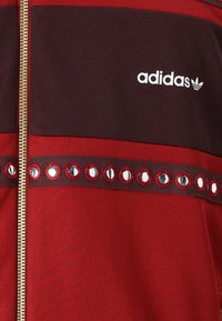 Röd och mörk marnjjacka med dragkedja som har glänsande runda nitar på ett horisontellt band, med vit "adidas"-logotyp ovanför dragkedjan.