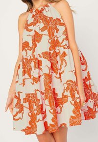 Ärmlös krämfärgad klänning med orange blommönster och samlade volanger, bärs av en kvinna som håller i sidorna av kjolen.