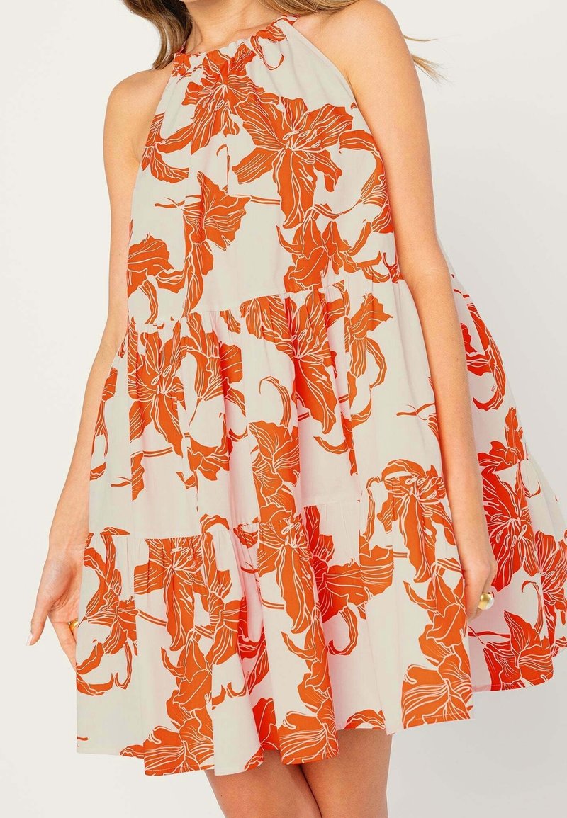 Ärmlös krämfärgad klänning med orange blommönster och samlade volanger, bärs av en kvinna som håller i sidorna av kjolen.