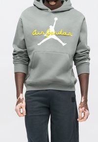 Personne portant un sweat à capuche Air Jordan gris avec un logo jaune et un pantalon foncé, debout avec un vitiligo visible sur les mains et les poignets.