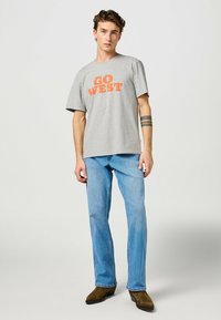 Camiseta de algodón gris con un gráfico naranja que dice "GO WEST", acompañada de jeans de mezclilla azul claro y calzado marrón. Diseño de corte relajado.