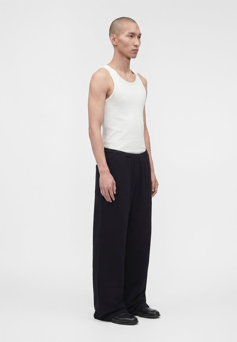 Homme portant un débardeur blanc sans manches, un pantalon noir ample et des chaussures noires, debout devant un fond gris clair uni.