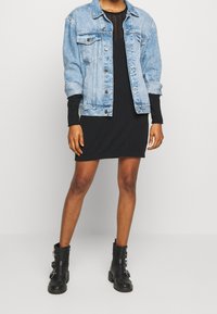 Persona che indossa un vestito nero, una giacca di jeans azzurro chiaro e stivali neri alla caviglia con fibbie, in piedi davanti a uno sfondo chiaro e neutro.