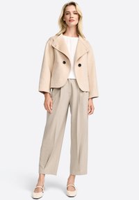 Giacca in misto lana beige cropped con grandi bottoni neri, abbinata a pantaloni leggermente plissettati grigi chiari e scarpe beige slip-on. Texture liscia.