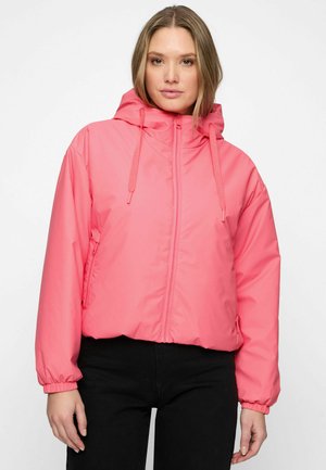 Vrouw draagt een felroze gewatteerde jas met capuchon, rits aan de voorkant en trekkoordjes, gecombineerd met een zwarte broek, staand tegen een witte achtergrond.