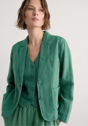Mujer con un chaleco verde abotonado y una blazer a juego con bolsillos, contra un fondo claro y liso.