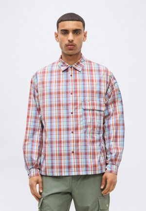 Jeune homme portant une chemise à carreaux rouge, bleue et blanche à manches longues avec une poche poitrine et un pantalon cargo vert, debout devant un fond uni.
