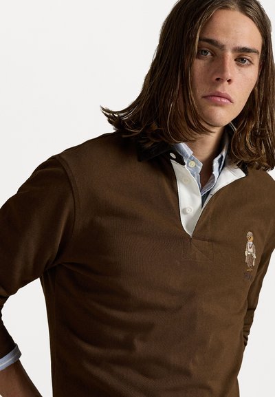 Bruine polo met lange mouwen, witte en blauwe kraag, met geborduurde karakterdetail op de borst en een textuurstof.
