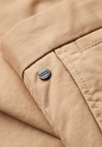 Tessuto di cotone marrone piegato, con un bottone nero con l'incisione "SUPERDRY PREMIUM". Superficie texturizzata con dettagli di cucitura visibili.