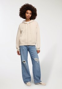 Felpa beige con cappuccio e tasche anteriori, abbinata a jeans a gamba larga chiari e strappati. Sneakers con un accento sottile completano l'outfit.