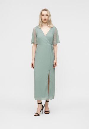 Frau trägt ein hellgrünes Wickelkleid mit kurzen Spitzärmeln und einem frontalen Schlitz, kombiniert mit schwarzen Sandalen mit Knöchelriemen und hohem Absatz.